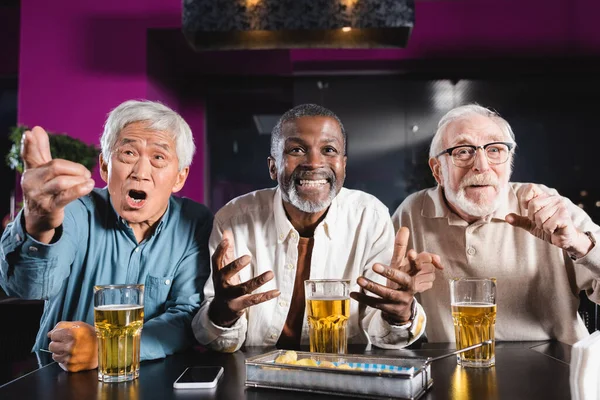 Emocionado amigos multiétnicos ancianos haciendo gestos mientras veía el partido de fútbol en el pub de cerveza - foto de stock