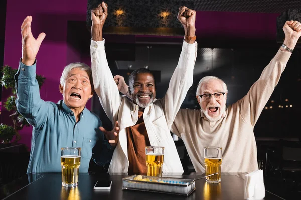 Excité amis multiethniques aînés regarder match de football dans le pub et montrant geste de victoire — Photo de stock