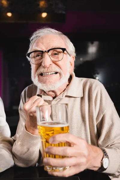 Worried senior man in eyeglasses holding blurred glass of beer while watching soccer championship in pub — Stock Photo