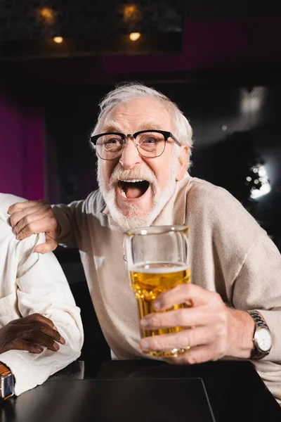 Excited senior man shouting and touching shoulder of african american friend while watching football in pub — Stock Photo