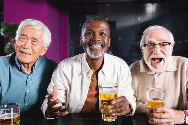 Thriller homens multiculturais seniores assistindo jogo de futebol perto de copos de cerveja no pub — Fotografia de Stock