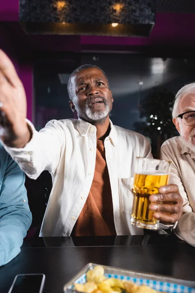 Chateado afro-americano homem apontando com a mão enquanto assiste jogo de futebol com amigos seniores no pub — Fotografia de Stock