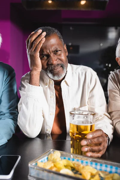 Bouleversé homme afro-américain avec un verre de bière toucher la tête tout en regardant le championnat dans le bar sportif — Photo de stock