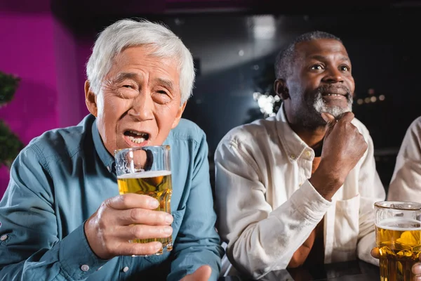 Preocupado asiático homem segurando copo de cerveja enquanto assiste futebol com interracial amigos no pub — Fotografia de Stock