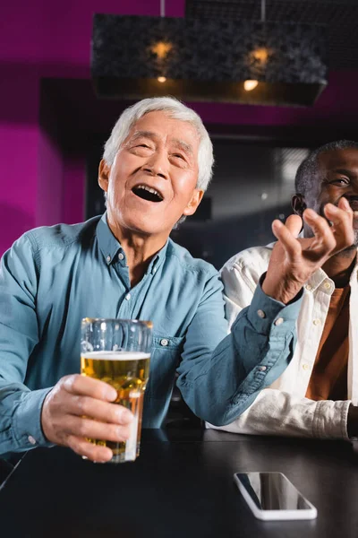 Erstaunt asiatischer Mann mit Glas Bier beobachtet Meisterschaft in Sportbar in der Nähe von Senioren multiethnischen Freunden — Stockfoto