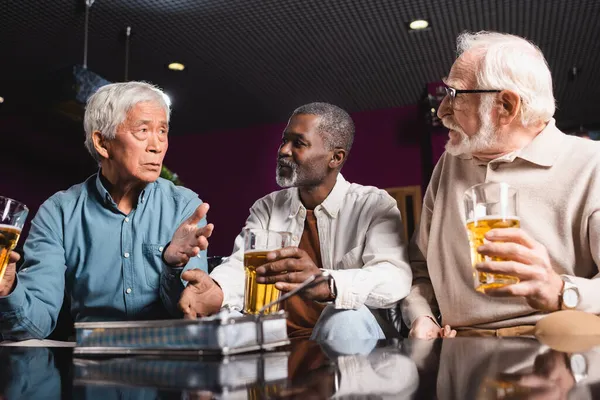 Amigos mayores haciendo gestos y sosteniendo vasos de cerveza durante la conversación en el pub - foto de stock