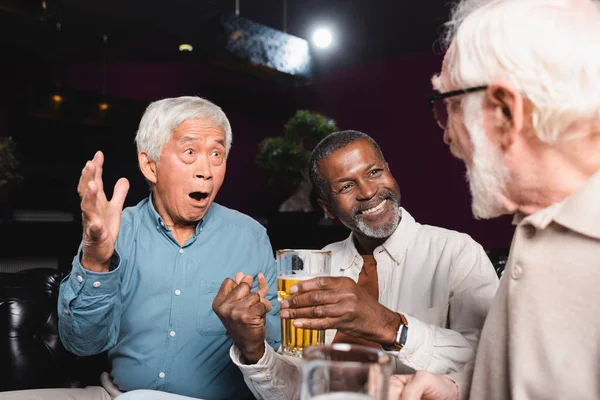 Asombrado asiático hombre gesto mientras hablando a ancianos multiétnicos amigos en cerveza pub - foto de stock