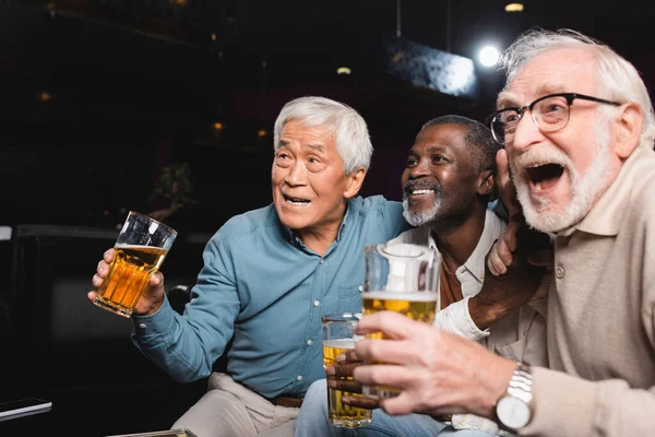 Amazed senior man with open mouth watching football in beer pub with happy interracial friends — Stock Photo