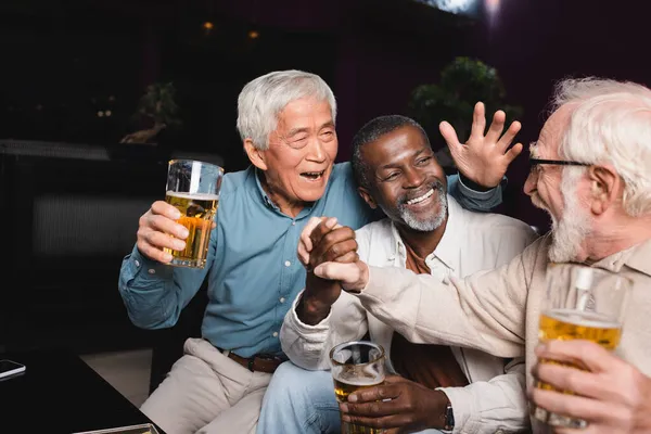 Amici multiculturali anziani e allegri che si uniscono le mani mentre tengono gli occhiali con la birra nel pub — Foto stock