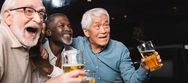 Hombre alegre gritando mientras ve el partido de fútbol con amigos mayores interracial en el pub, pancarta - foto de stock