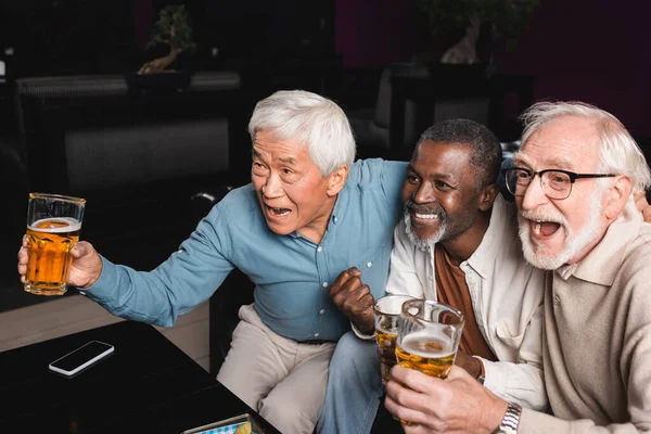 Sonriente afroamericano hombre con el puño cerrado mostrando partido de fútbol con emocionados amigos interracial en el pub - foto de stock