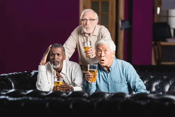 Sorprendió a amigos multiétnicos mayores con vasos de cerveza viendo el campeonato de fútbol en el pub - foto de stock