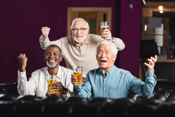 Idosos alegres amigos multiétnicos segurando cerveja e mostrando gesto de vitória enquanto assiste jogo de futebol no bar — Fotografia de Stock