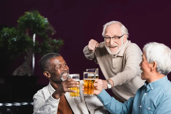 Emocionado hombre mayor mostrando gesto de victoria mientras tintineo vasos de cerveza con amigos en el pub - foto de stock