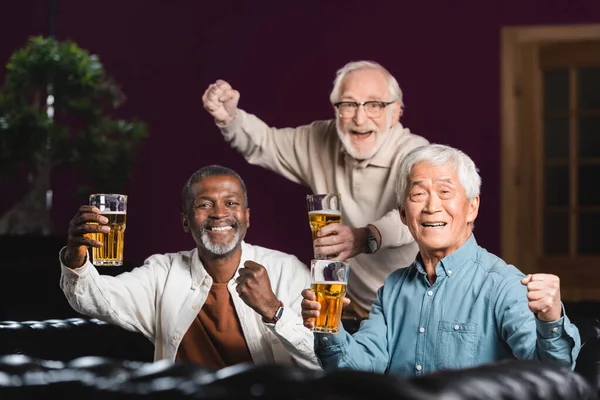 Amigos multiculturais alegres com copos de cerveja mostrando gesto de vitória no bar de esportes — Fotografia de Stock