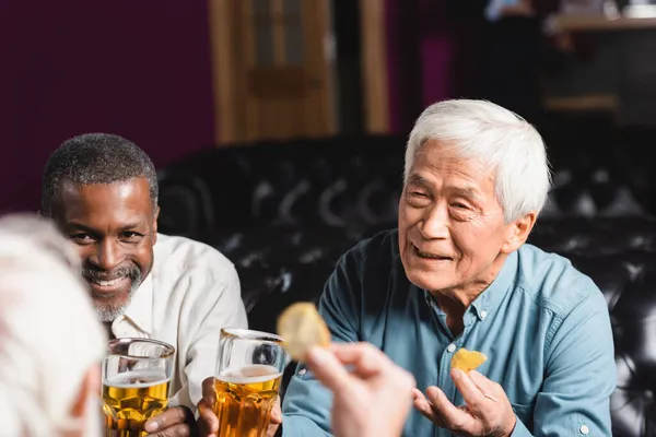 Senior multicultural friends talking, drinking beer and eating chips in pub — Stock Photo