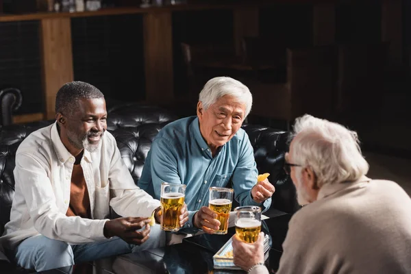 Amigos multiculturales mayores sosteniendo vasos de cerveza mientras pasan tiempo en el pub - foto de stock