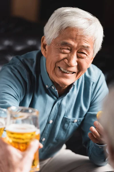 Alegre senior asiático hombre tintineo vasos de cerveza con borrosa amigo en pub - foto de stock