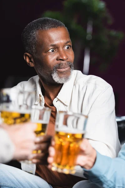 Изумленный пожилой афроамериканец, звенящий размытыми пивными бокалами с друзьями в пабе — стоковое фото
