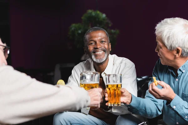 Fröhlicher afrikanisch-amerikanischer Mann blickt in die Kamera, während er mit verschwommenen älteren Freunden Biergläser klirrt — Stockfoto