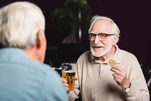 Sonriente anciano sosteniendo un vaso de cerveza y patatas fritas cerca de un amigo borroso en el pub - foto de stock