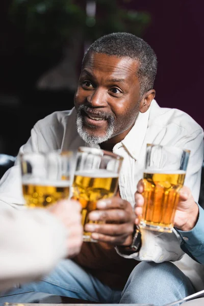 Älterer afrikanisch-amerikanischer Mann lächelt, während er mit Freunden verschwommene Biergläser klimpert — Stockfoto