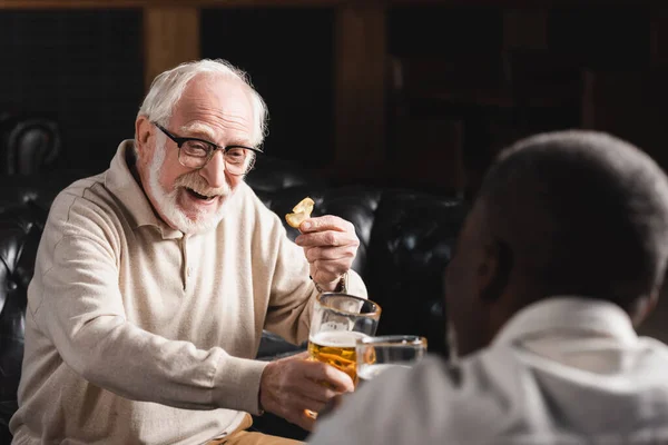 Feliz hombre mayor tintineo vasos de cerveza con borrosa africano americano amigo - foto de stock