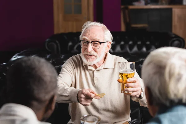 Homme âgé joyeux tenant des chips et de la bière tout en parlant à des amis interracial flous dans le pub — Photo de stock