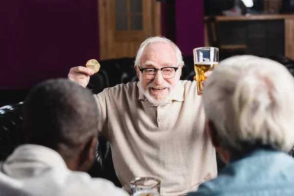 Animado homem sênior com cerveja e batatas fritas falando com amigos multiétnicos desfocados no pub — Fotografia de Stock