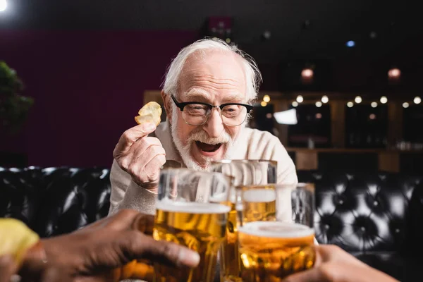 Excited senior man holding chips while clinking beer glasses with blurred interracial friends — Stock Photo