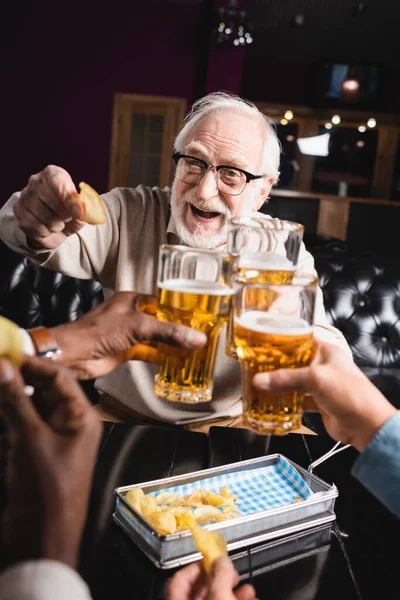 Cheerful elderly man holding chips while clinking beer glasses with blurred friends — Stock Photo