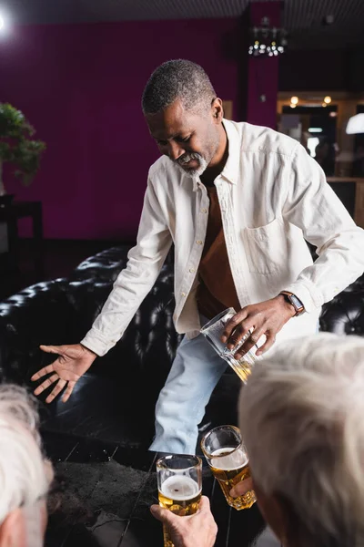 Senior afrikanisch-amerikanischer Mann hält ein Glas Bier in der Hand, während er neben verschwommenen Freunden in der Kneipe steht — Stockfoto