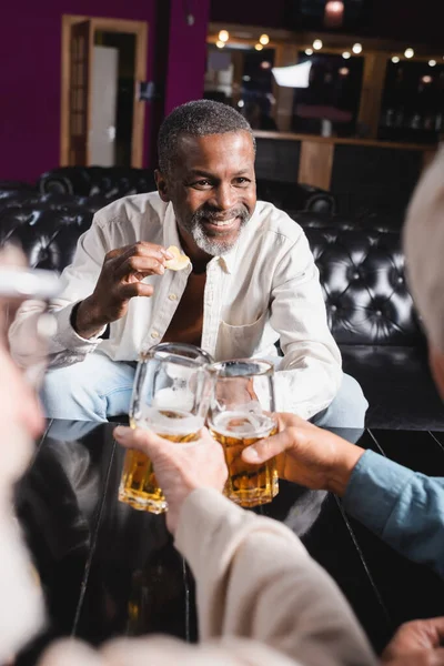 Feliz homem americano africano sênior segurando batatas fritas enquanto clinking copos de cerveja com amigos borrados — Fotografia de Stock