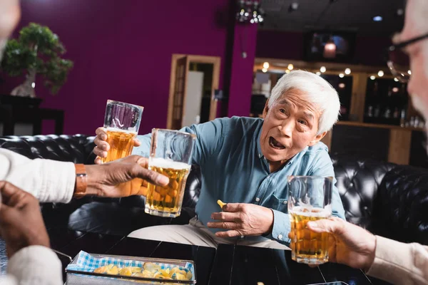 Возбужденный старший азиатский мужчина тост с пивом рядом размытые многоэтнических друзей в пабе — Stock Photo