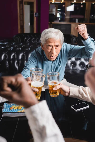 Excited senior asian man showing win gesture while clinking beer glasses with blurred interracial friends — Stock Photo