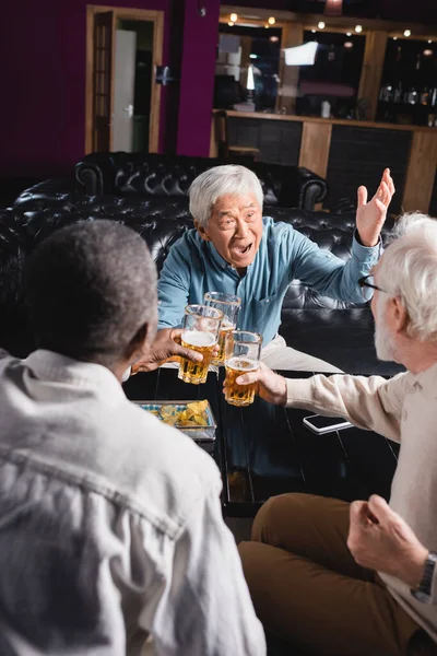 Thrilled asian man gesturing while clinking glasses of beer with senior interracial friends in pub — Stock Photo