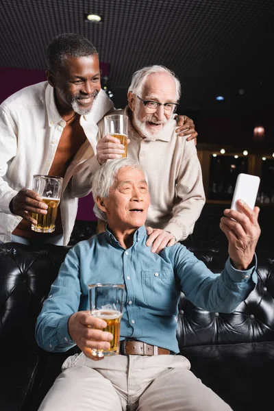 Senior asiatique homme assis sur canapé et prendre selfie avec des amis âgés dans bière pub — Photo de stock