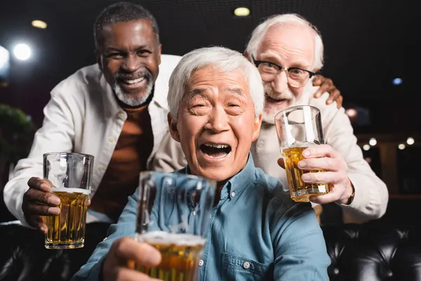 Alegres amigos multiétnicos sosteniendo vasos con cerveza mientras miran a la cámara en el pub - foto de stock