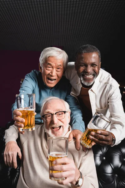 Excited, senior multicultural friends with glasses of beer looking at camera in bar — Stock Photo