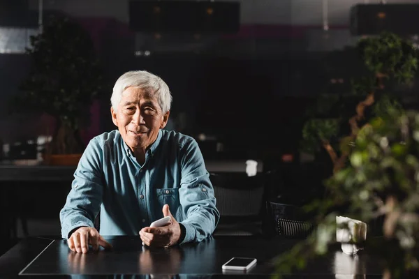 Старший азиатский мужчина держит смартфон и улыбается в камеру, сидя в кафе — стоковое фото