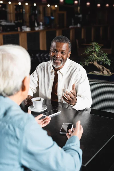 Happy african american man talking to blurred senior friend near smartphones with blank screen in cafe — Stock Photo