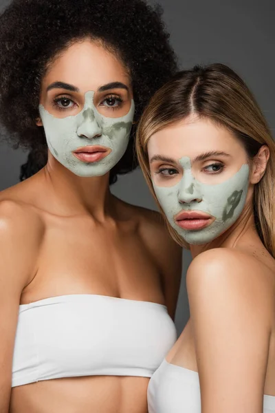 Jóvenes mujeres multiétnicas mimando caras con máscaras de arcilla aisladas en gris - foto de stock