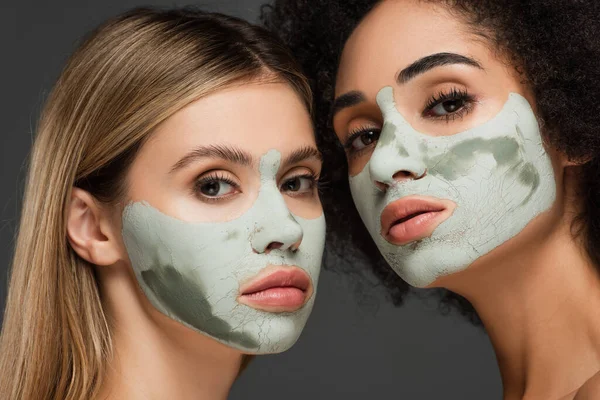 Close up view of multiethnic women in clay masks looking at camera isolated on grey — Stock Photo