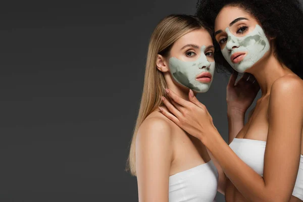 Multiethnic woman in white tops and clay masks posing isolated on grey — Stock Photo