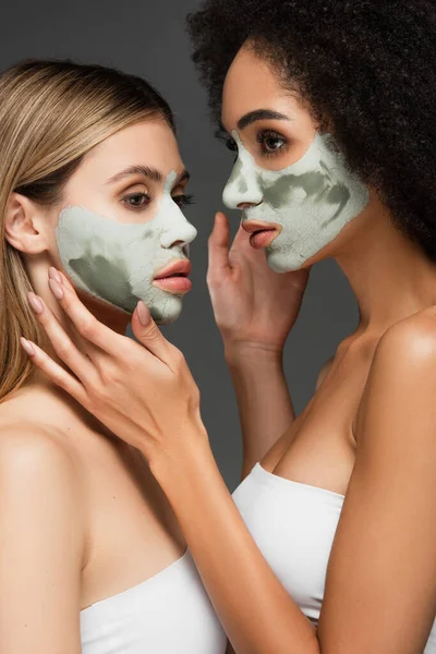 Multiethnic women in facial masks touching faces of each other isolated on grey — Stock Photo