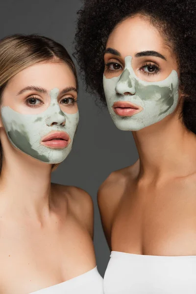 Young multiethnic women looking at camera while pampering faces with clay masks isolated on grey — Stock Photo