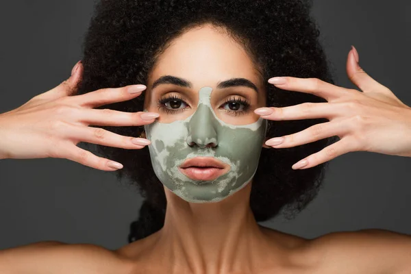 Donna afroamericana con maschera di argilla e le mani vicino al viso guardando la fotocamera isolata sul grigio — Foto stock