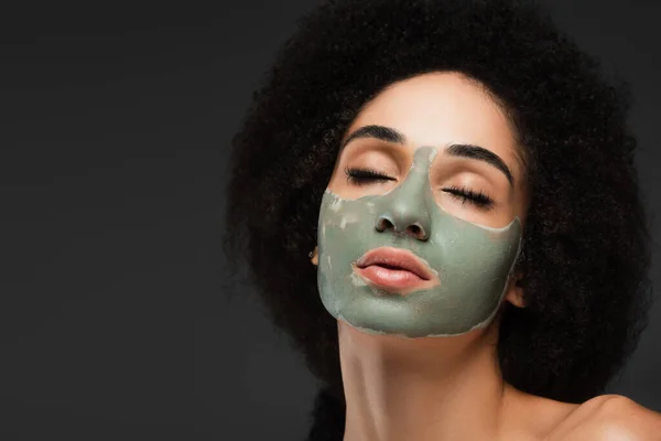 Young african american woman with closed eyes pampering face with clay mask isolated on grey — Stock Photo