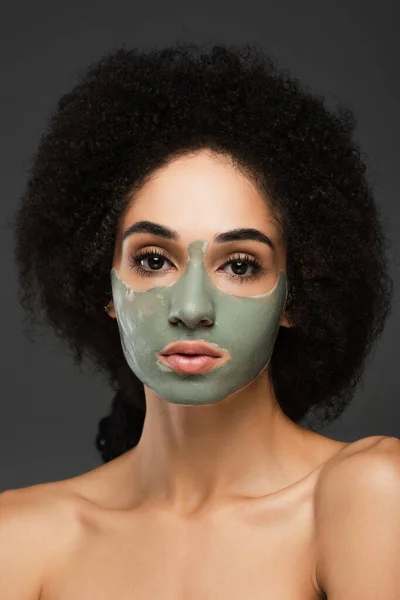 Pretty african american woman with naked shoulders and clay facial mask looking at camera isolated on grey — Stock Photo