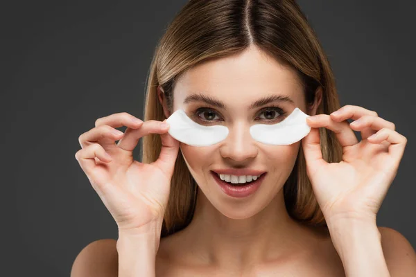 Femme souriante regardant la caméra tout en tenant les patchs pour les yeux isolés sur gris — Photo de stock